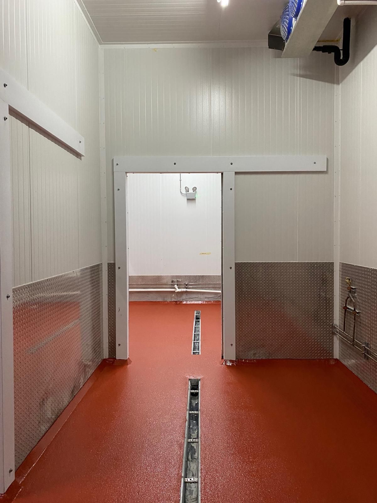 Narrow industrial hallway with red floor, white walls, and a central doorway with a clear strip drain.