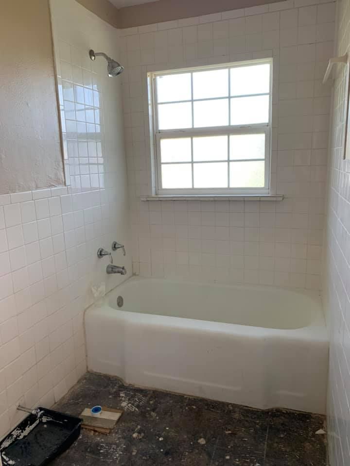 White-tiled bathroom with a bathtub, shower, and window. The floor has paint splatters and a paint tray.