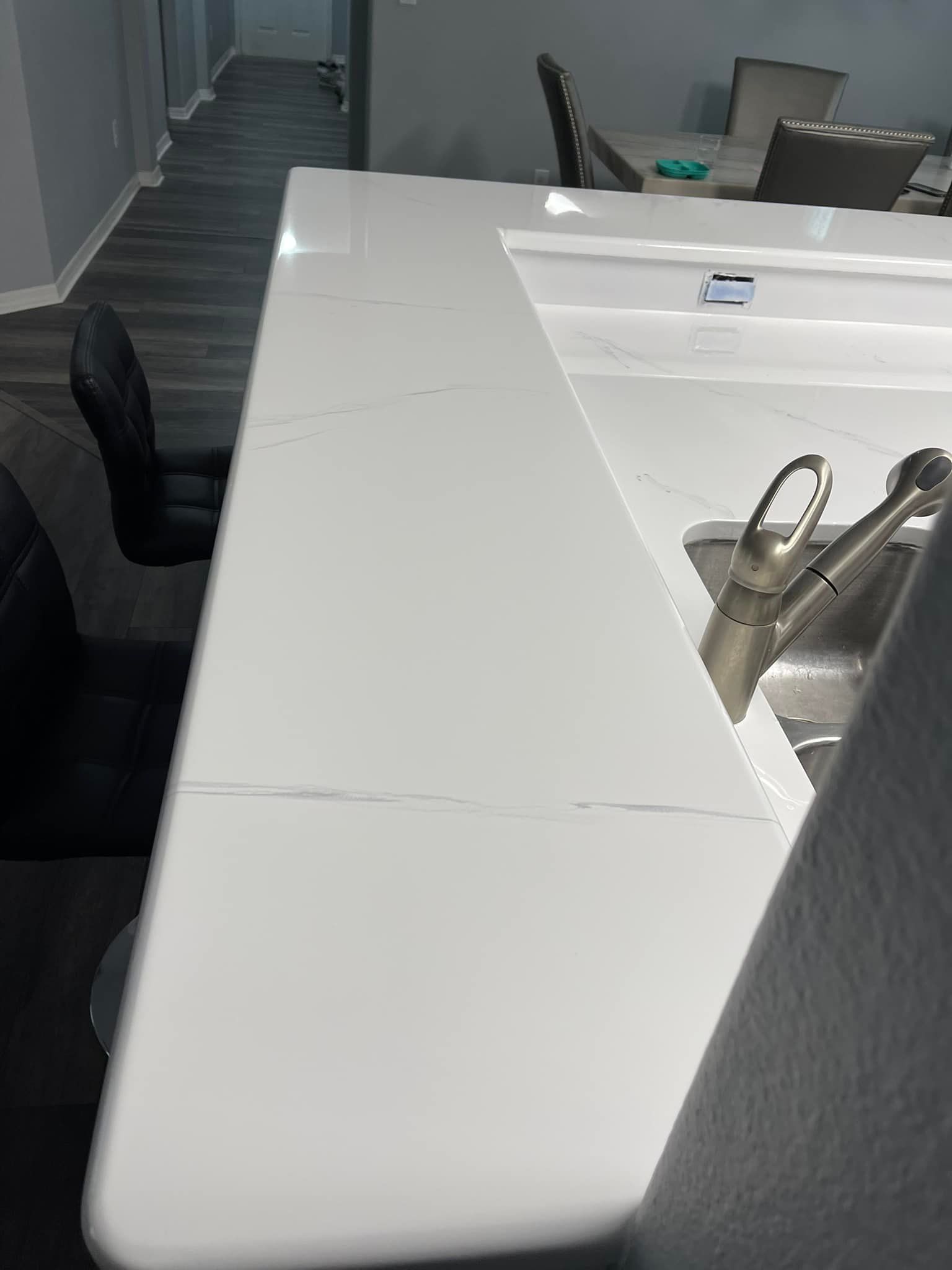 White kitchen countertop with a stainless steel sink and faucet. Black barstool in the foreground.