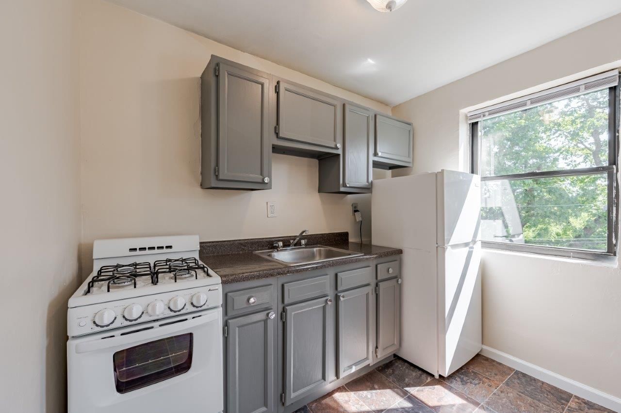 Small kitchen with gray cabinets, white stove and fridge, and a window letting in daylight.