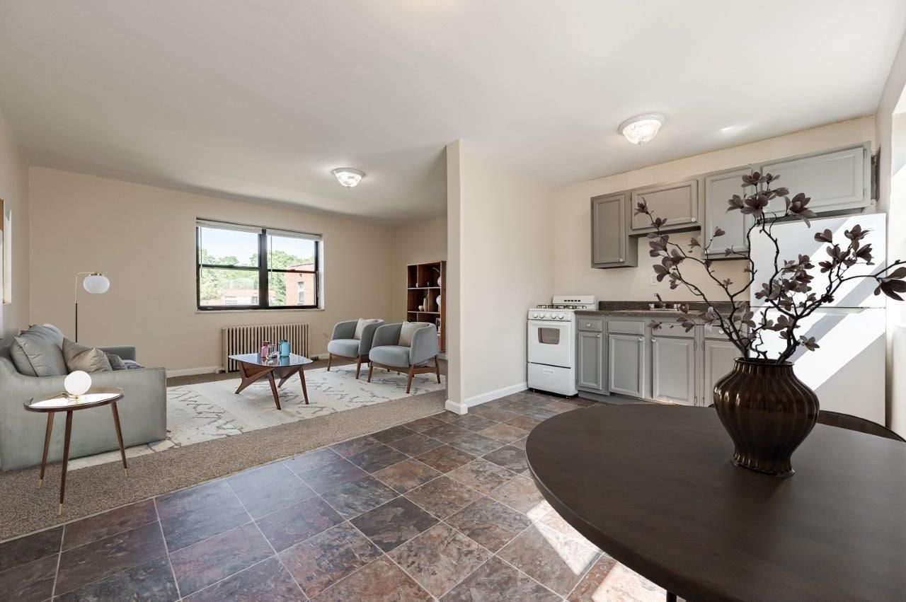 Open-concept living room and kitchenette with tile floors, gray seating, and a round table with flowers.