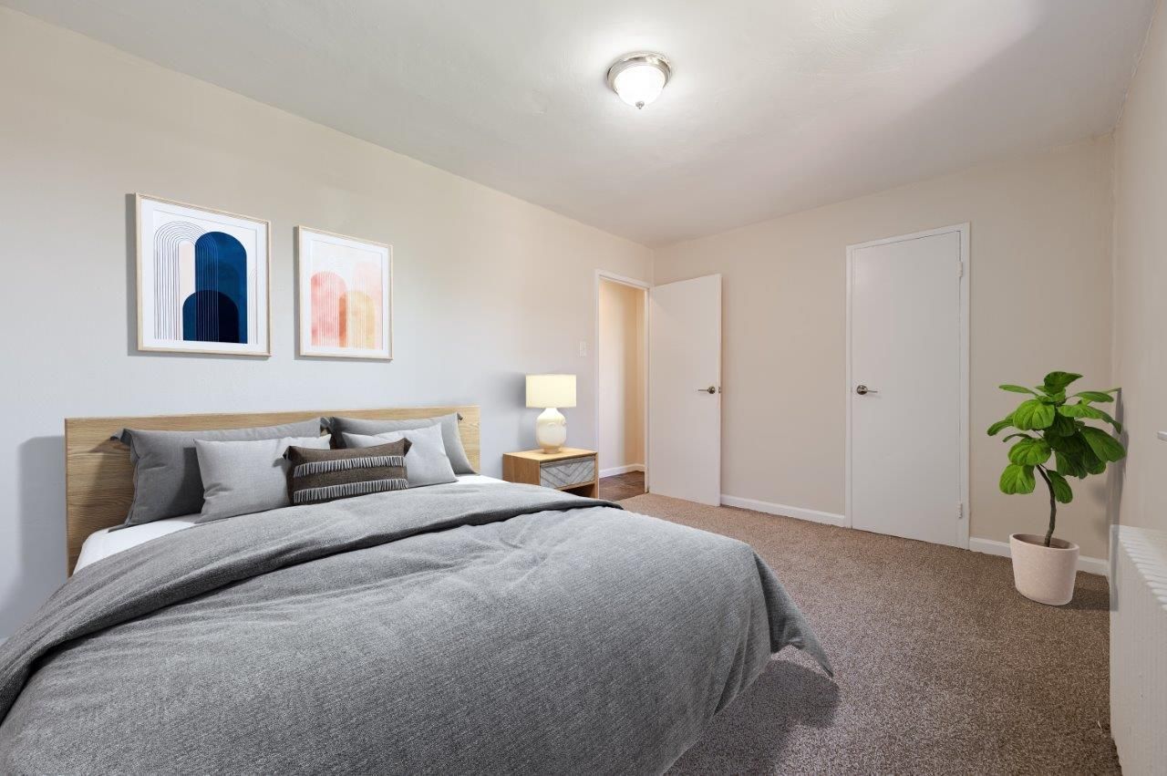 Cozy bedroom with gray bed, nightstand lamp, wall art, carpet, closet doors, and a potted plant.
