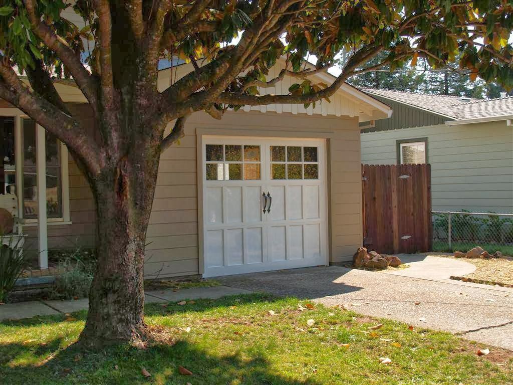 Suburban house with a white garage door, large tree in front, and a driveway beside a wooden fence.