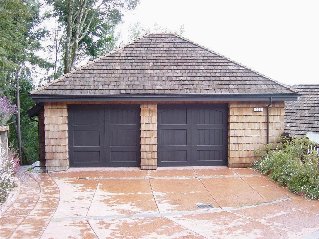 Stone garage with two dark doors and a shingled roof beside a wet driveway and garden.