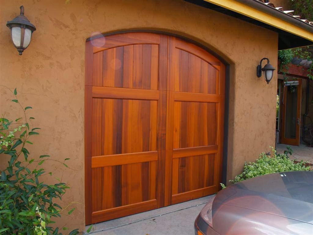 Wooden arched garage door on a tan stucco house, with wall lanterns and a car parked nearby