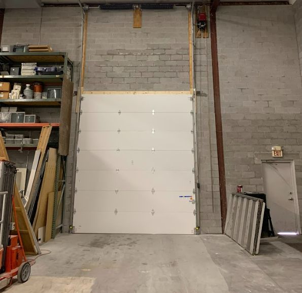 Warehouse interior with a large white roll-up garage door centered on a concrete floor, shelves on the left.