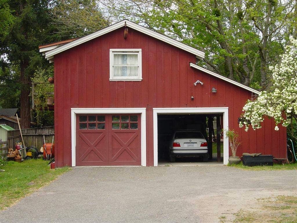 Red barn-style garage with a car parked inside and a white-trimmed upper window, surrounded by trees.