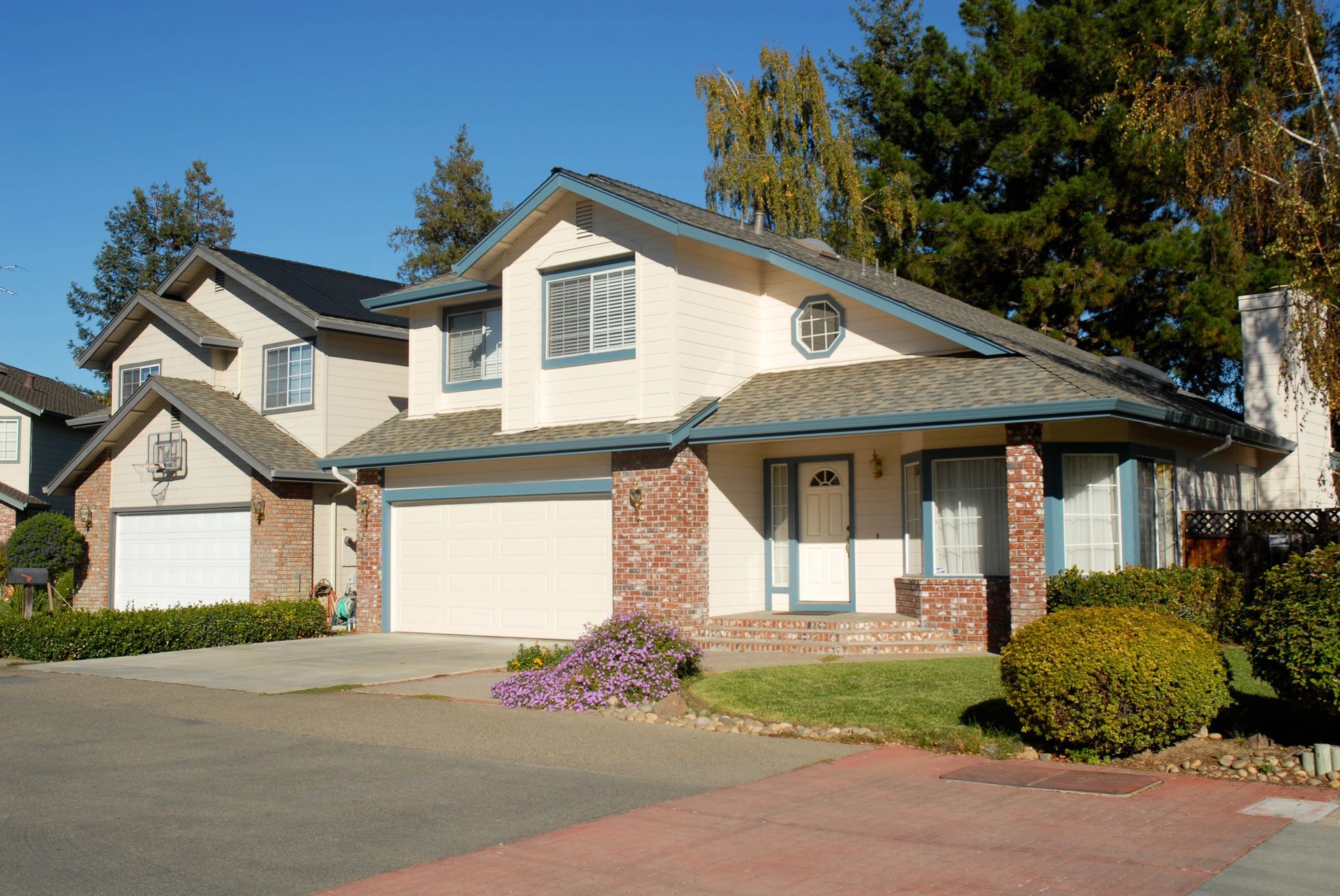 Suburban two-story house with beige siding, brick accents, white garage doors, and a landscaped front yard