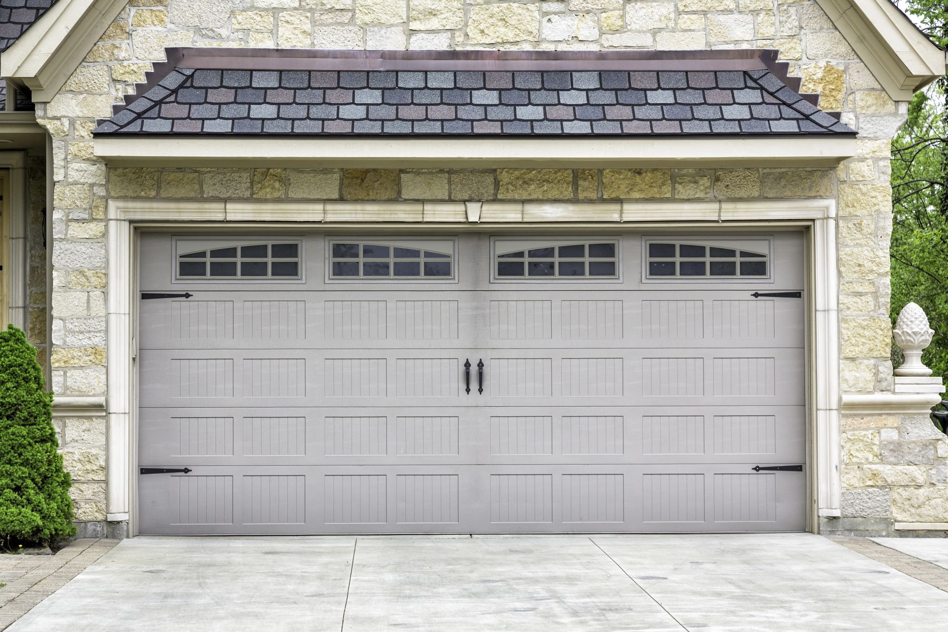 Stone house garage with a closed light gray double door and dark shingled roof trim