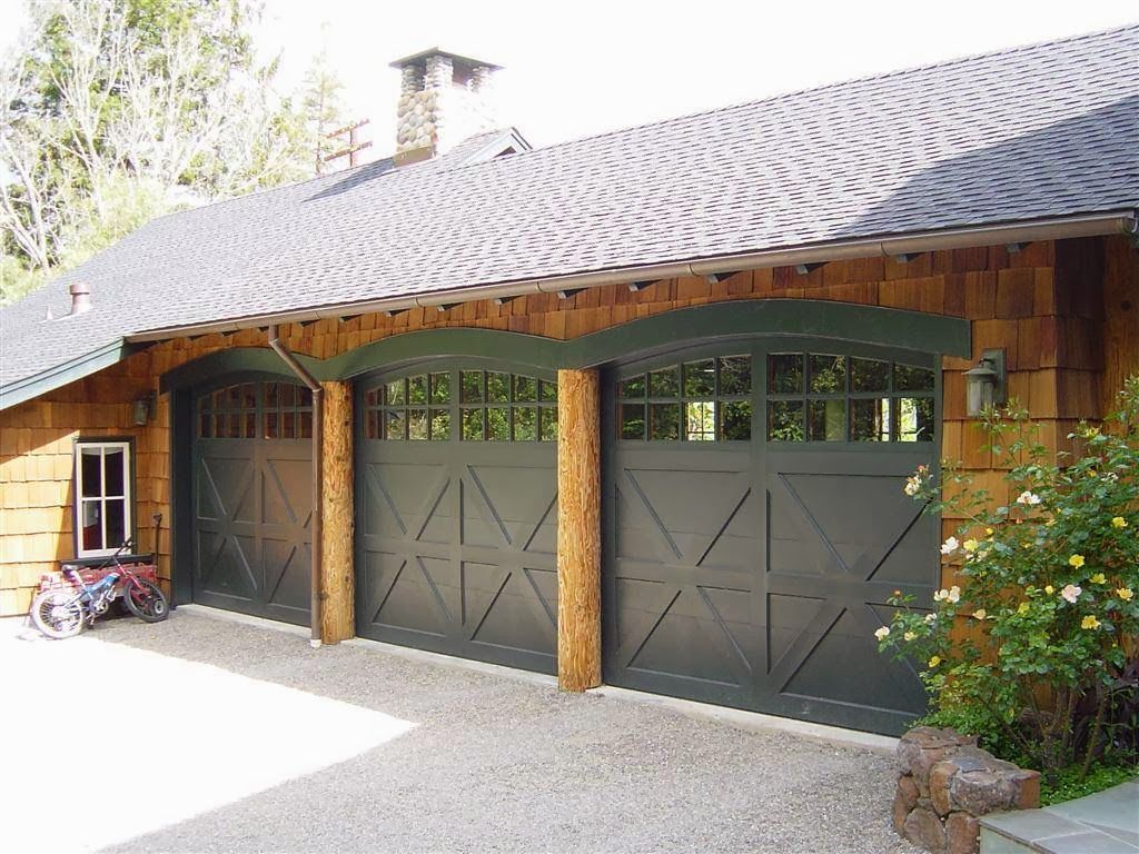 Three-car garage with gray doors, stone pillars, and a shingled roof beside greenery