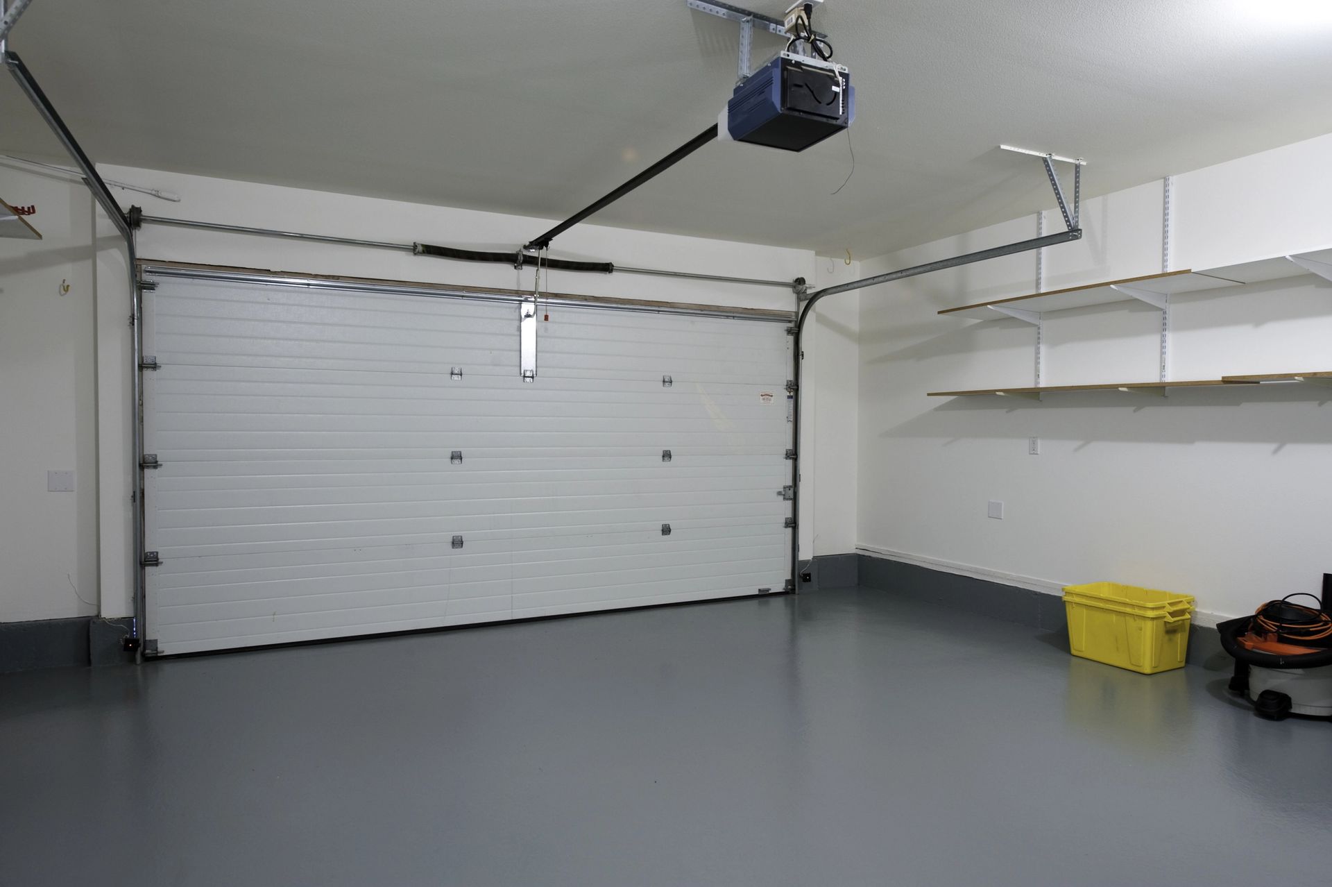 Empty garage with white walls, gray floor, overhead door, shelves, and a yellow storage bin.