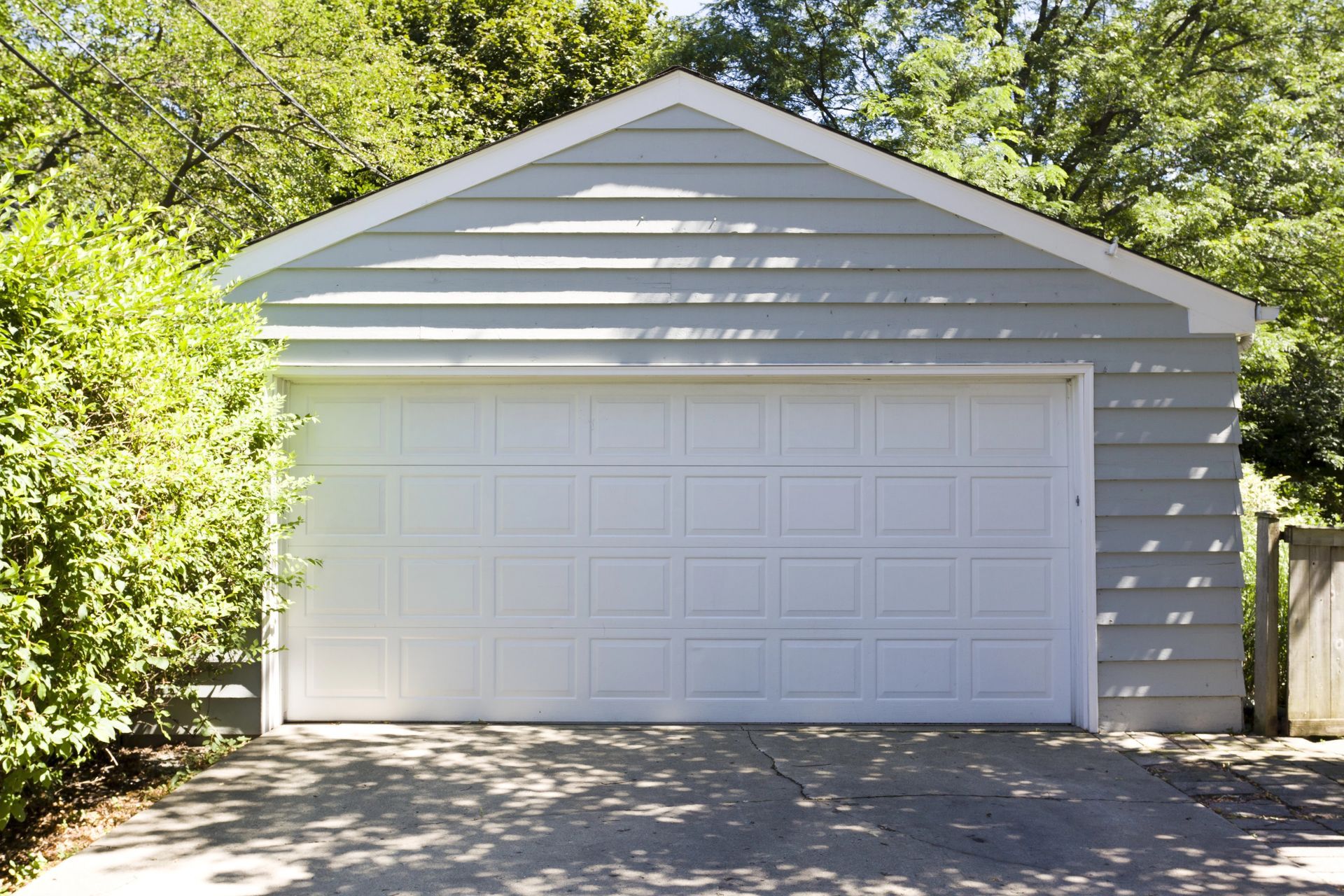 White detached garage with a paneled door, framed by green trees and driveway shadows