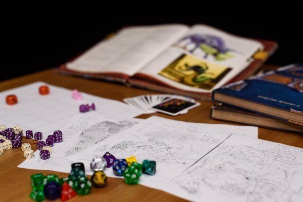 A tabletop gaming scene: rule books, dice, game pieces, and maps on a wooden surface.