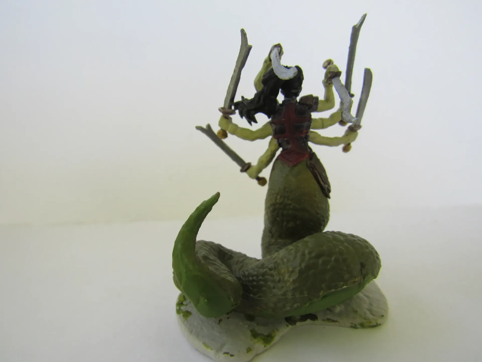 A multi-armed, snake-bodied figure holding swords. Dark hair, red chest, green scales, on a stone base.