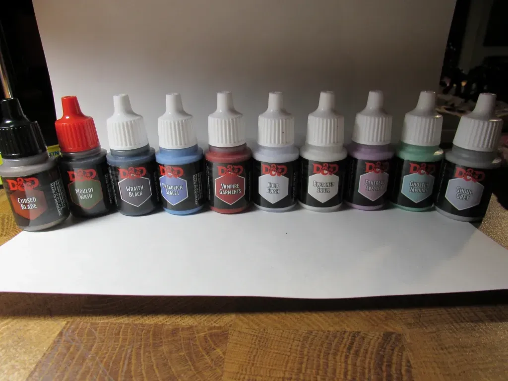 Ten bottles of D&D ink paints in various colors, lined up on a white surface, with a wooden table in the foreground.