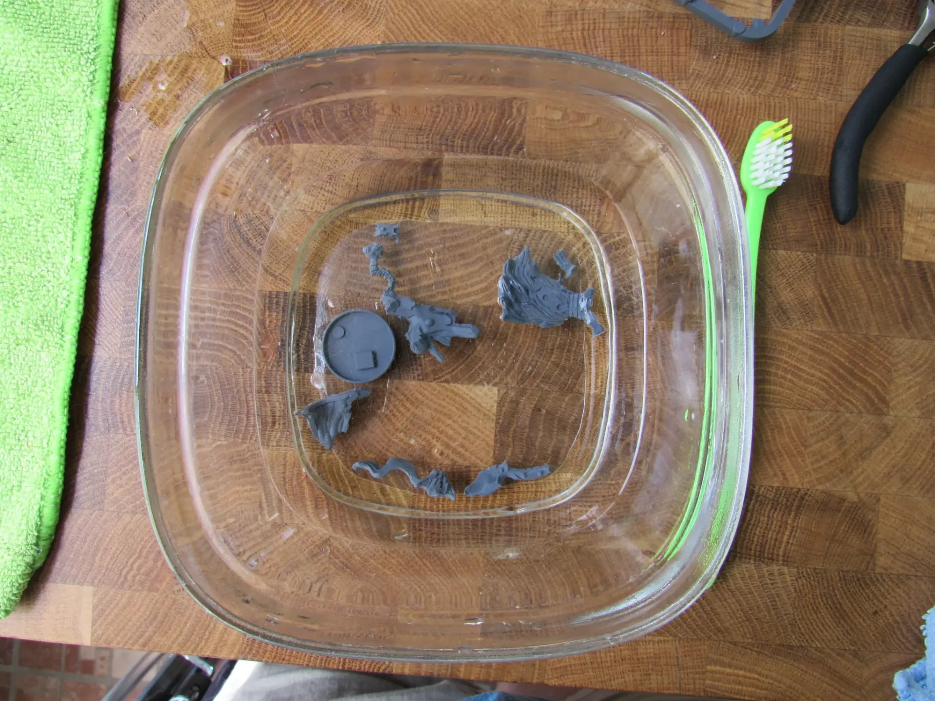 A glass dish with miniature figurines soaking in liquid, a toothbrush, and green cloth on a wooden surface.