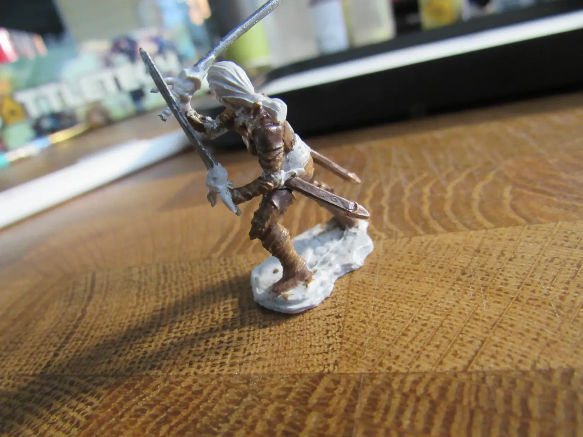 Miniature figure of a warrior with two swords in brown armor on a stone base.
