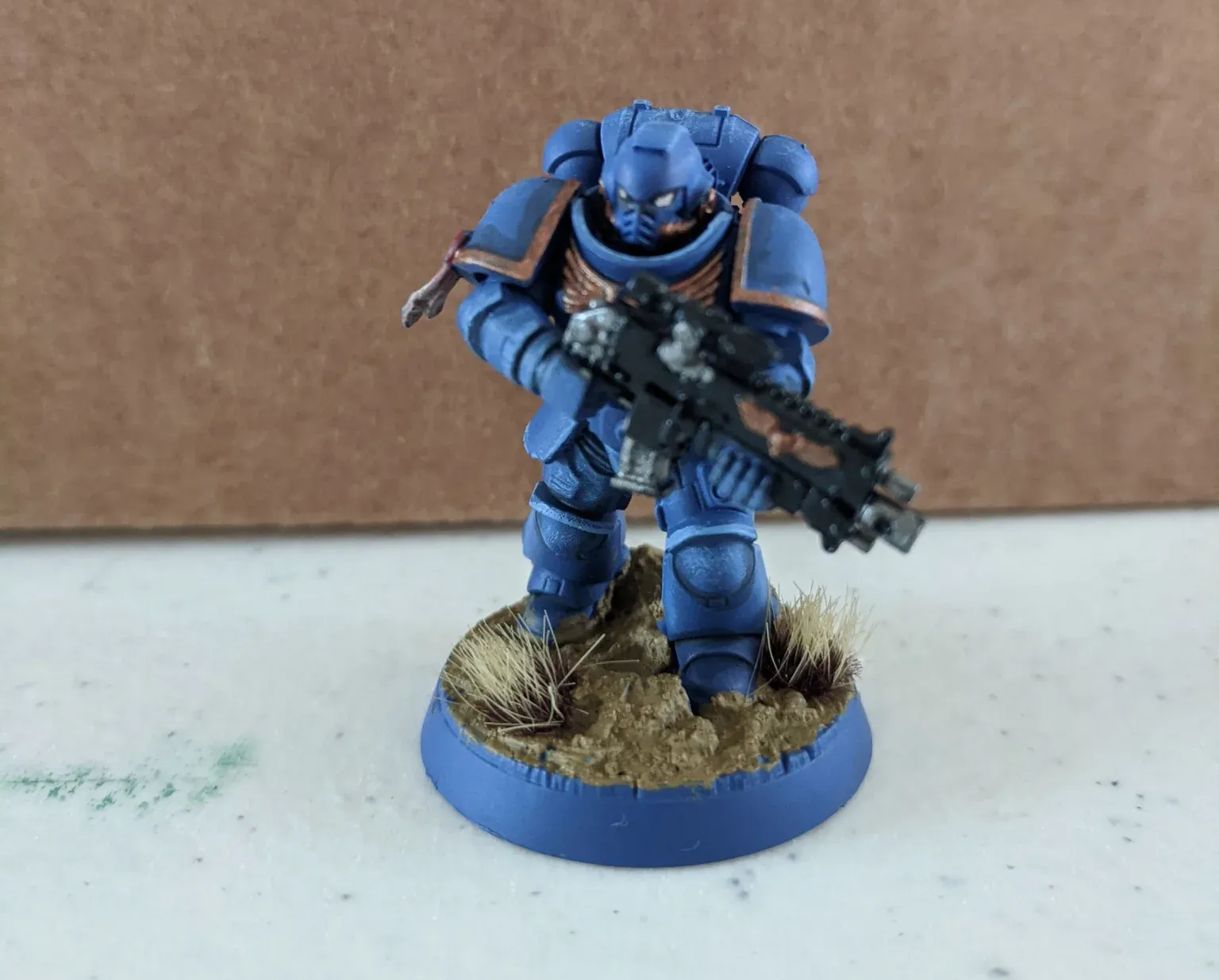 A blue miniature Space Marine soldier stands on a circular base. He holds a black gun, and the base has brown terrain.