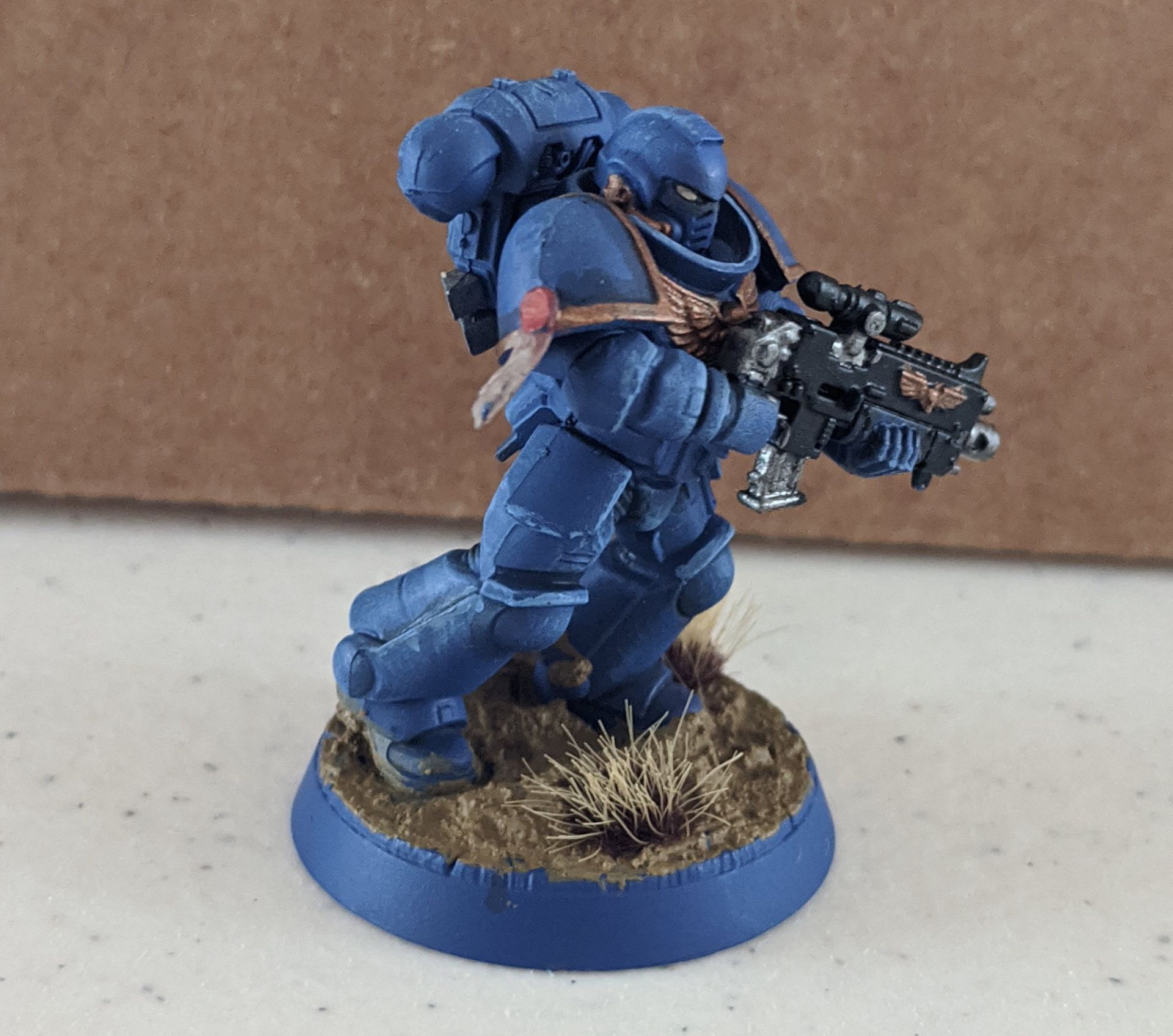 Blue-armored Space Marine miniature with a bolter, kneeling on a brown base, against a beige background.