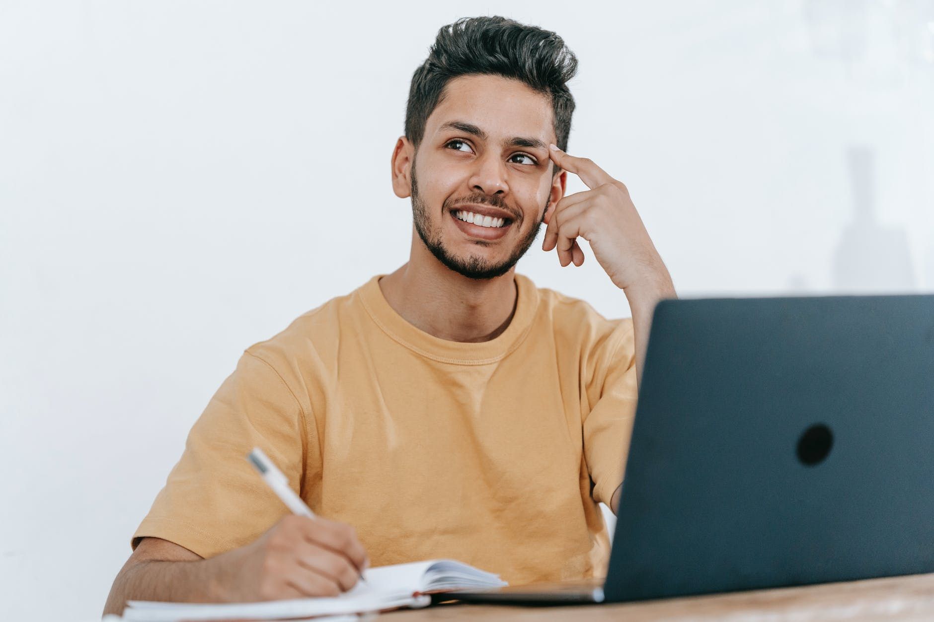 Man smiles while brainstorming, writing in a notebook next to an open laptop. He wears a yellow shirt.
