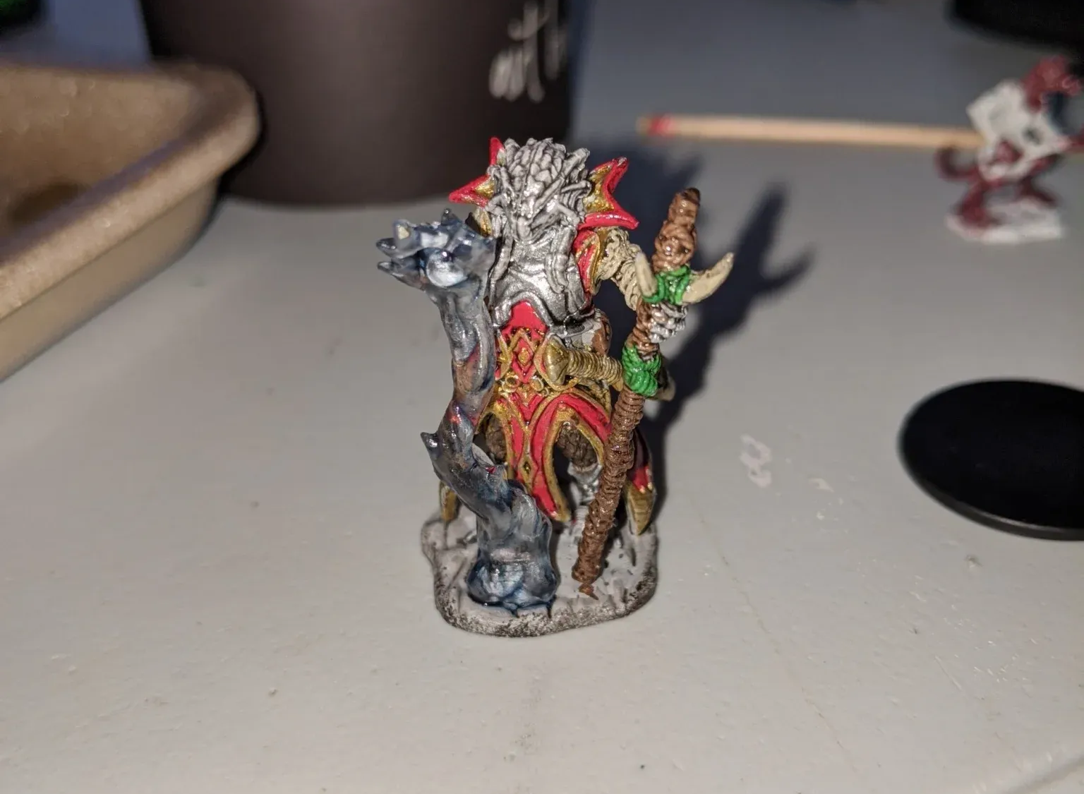 Miniature figure of a warrior in red and gold armor, on a black base, tabletop setting.