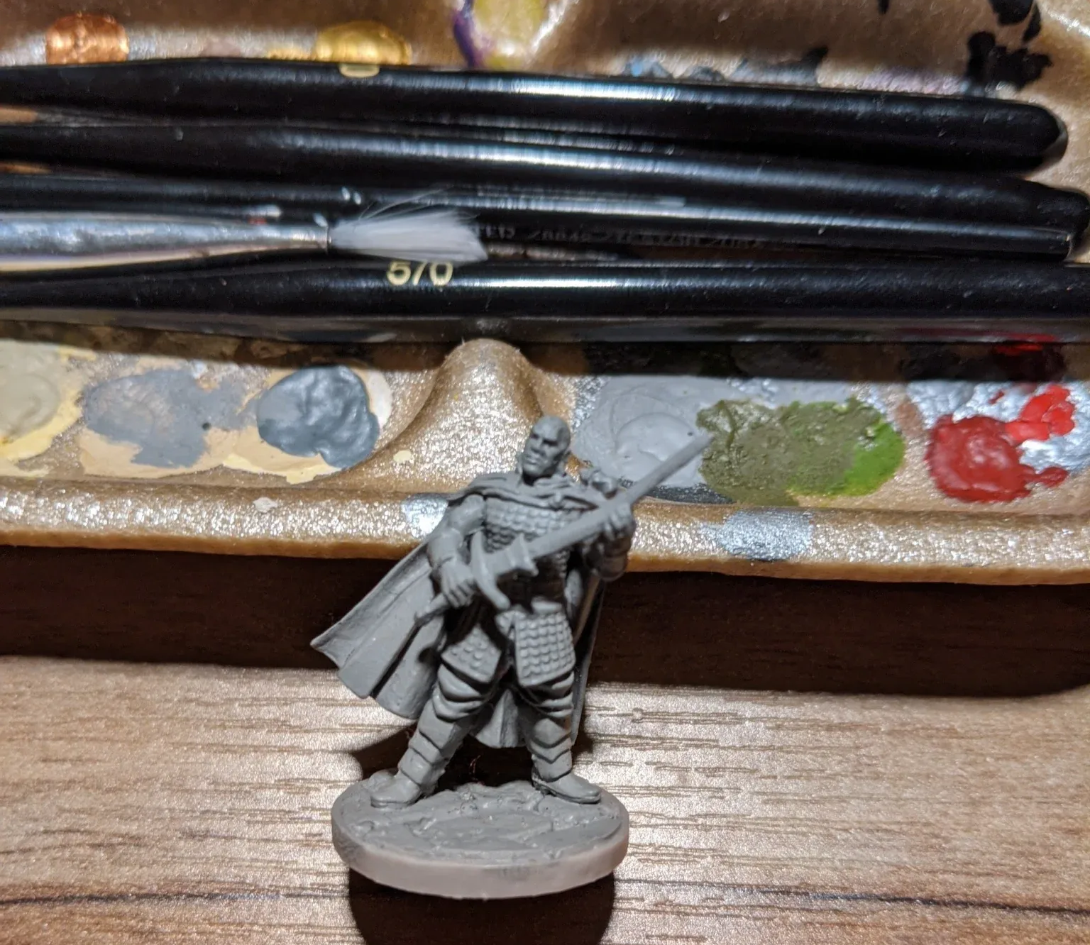 Miniature figure of a warrior in armor, holding a sword, with brushes and a paint palette in the background.