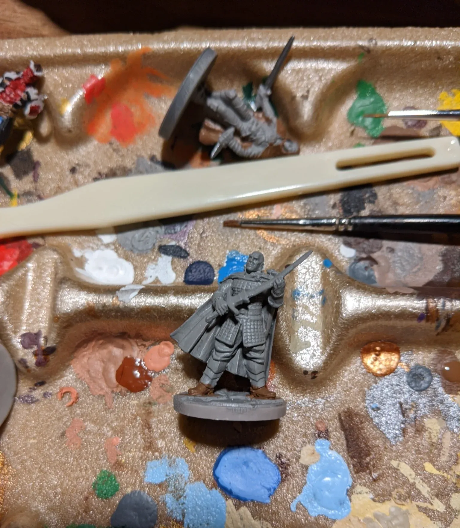 Miniature figure being painted on a palette. It features a figure holding a rifle and other figures. Paint and tools visible.
