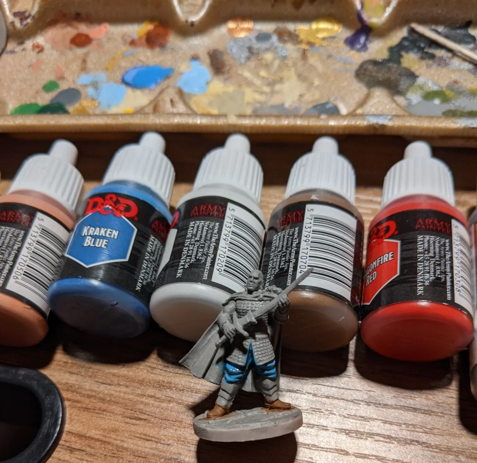 Miniature figure with blue, white, and brown paint next to a palette of colors and paint bottles.
