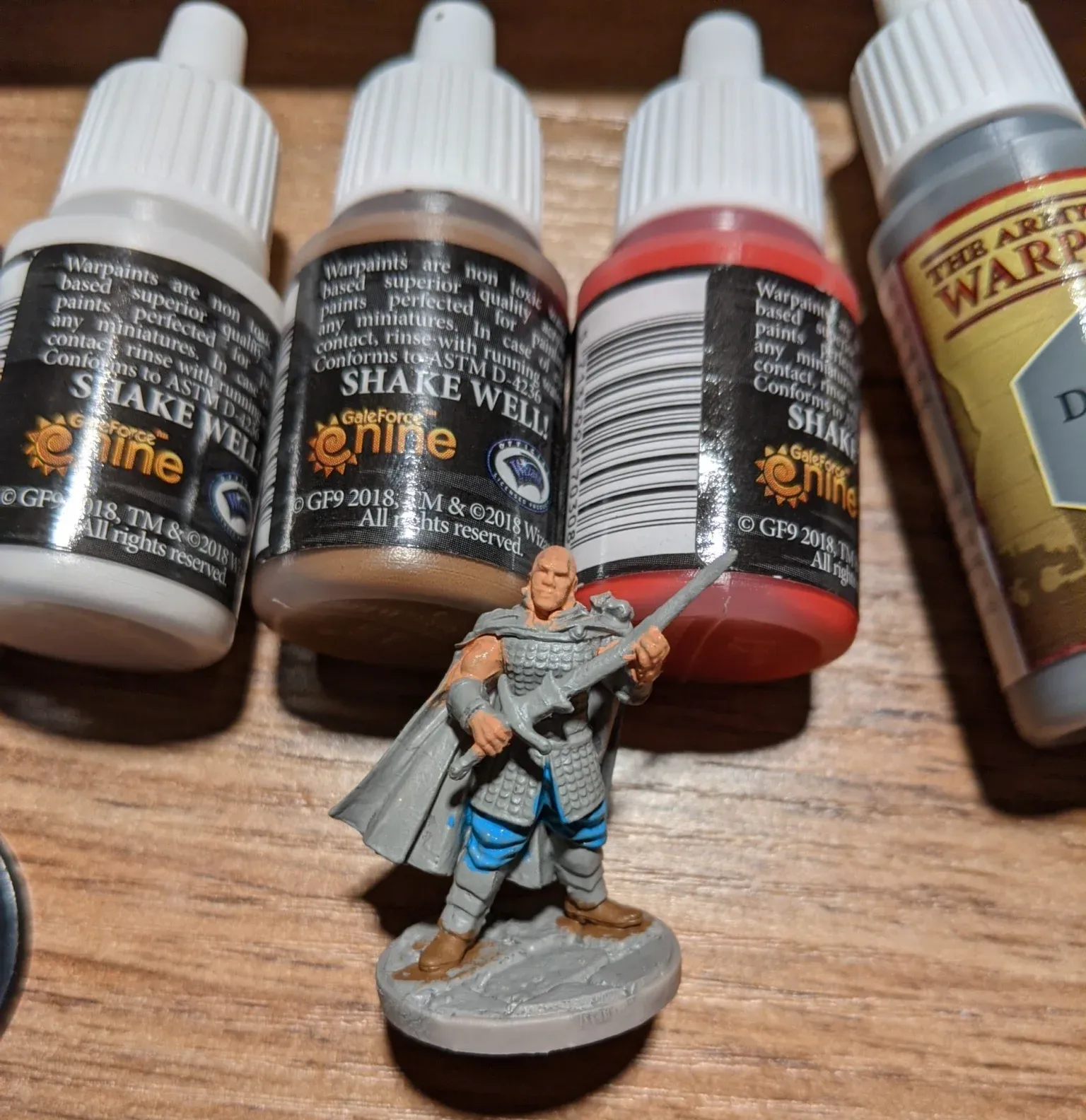 Miniature figure with paint bottles, depicting a bald, muscular man in armor, holding a sword on a rocky base.