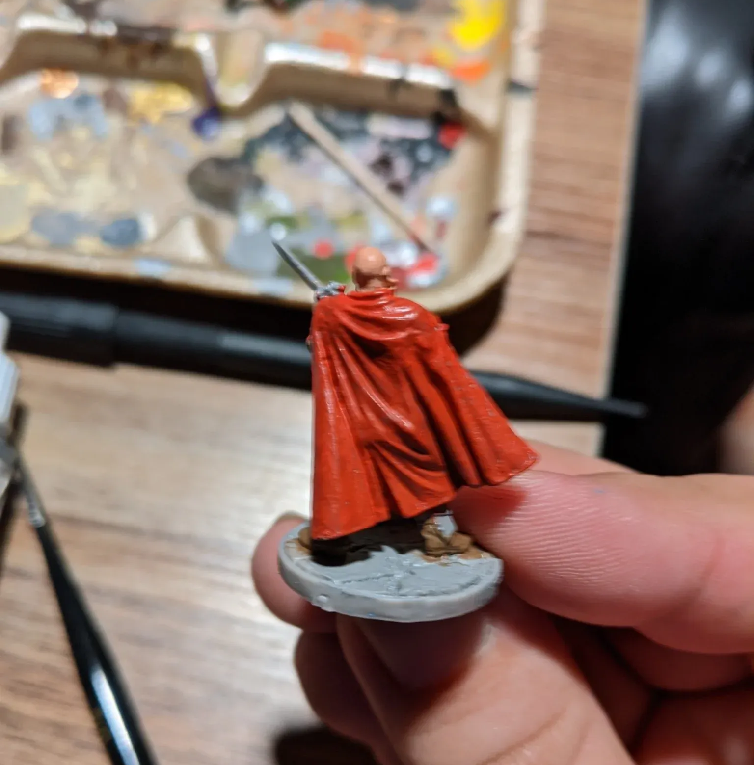 Miniature figurine of a bald figure in a red cape, holding a sword. Held in a hand.