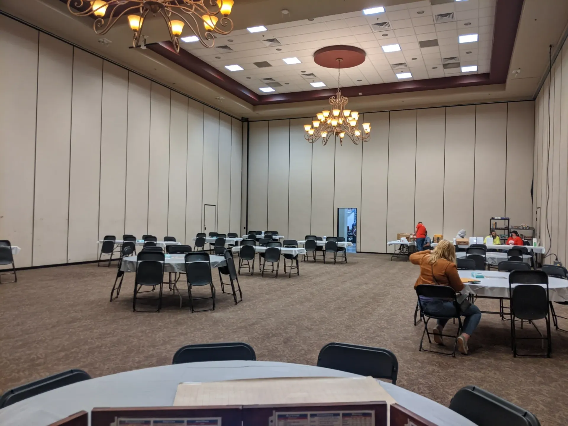 A large, empty banquet hall with tables, chairs, and chandeliers. People prepare in the background.
