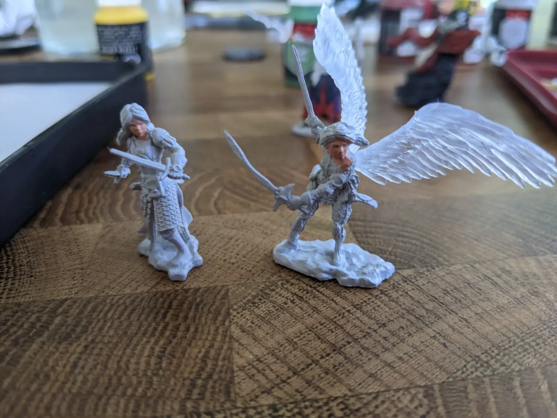 Two unpainted miniature fantasy figures on a wooden table. One is a person, the other winged with a sword.