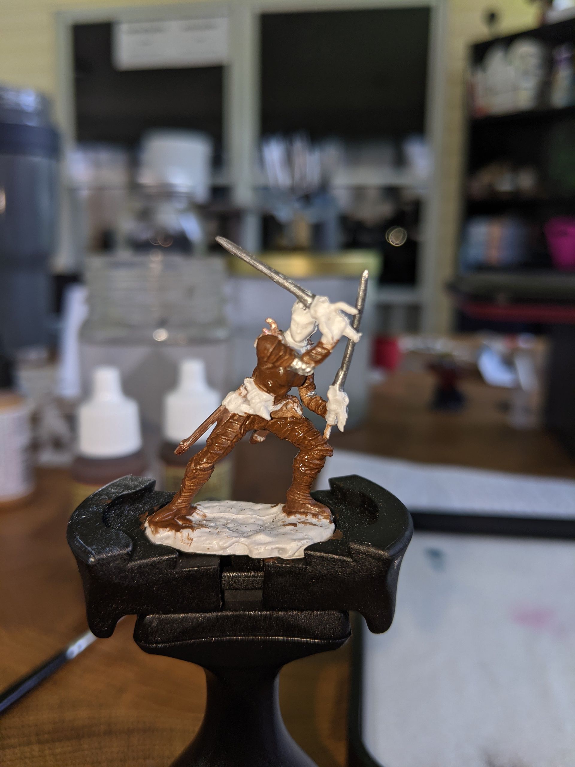 Miniature figure of a warrior, brown base, white and brown paint on the figure, poised with swords, indoors.
