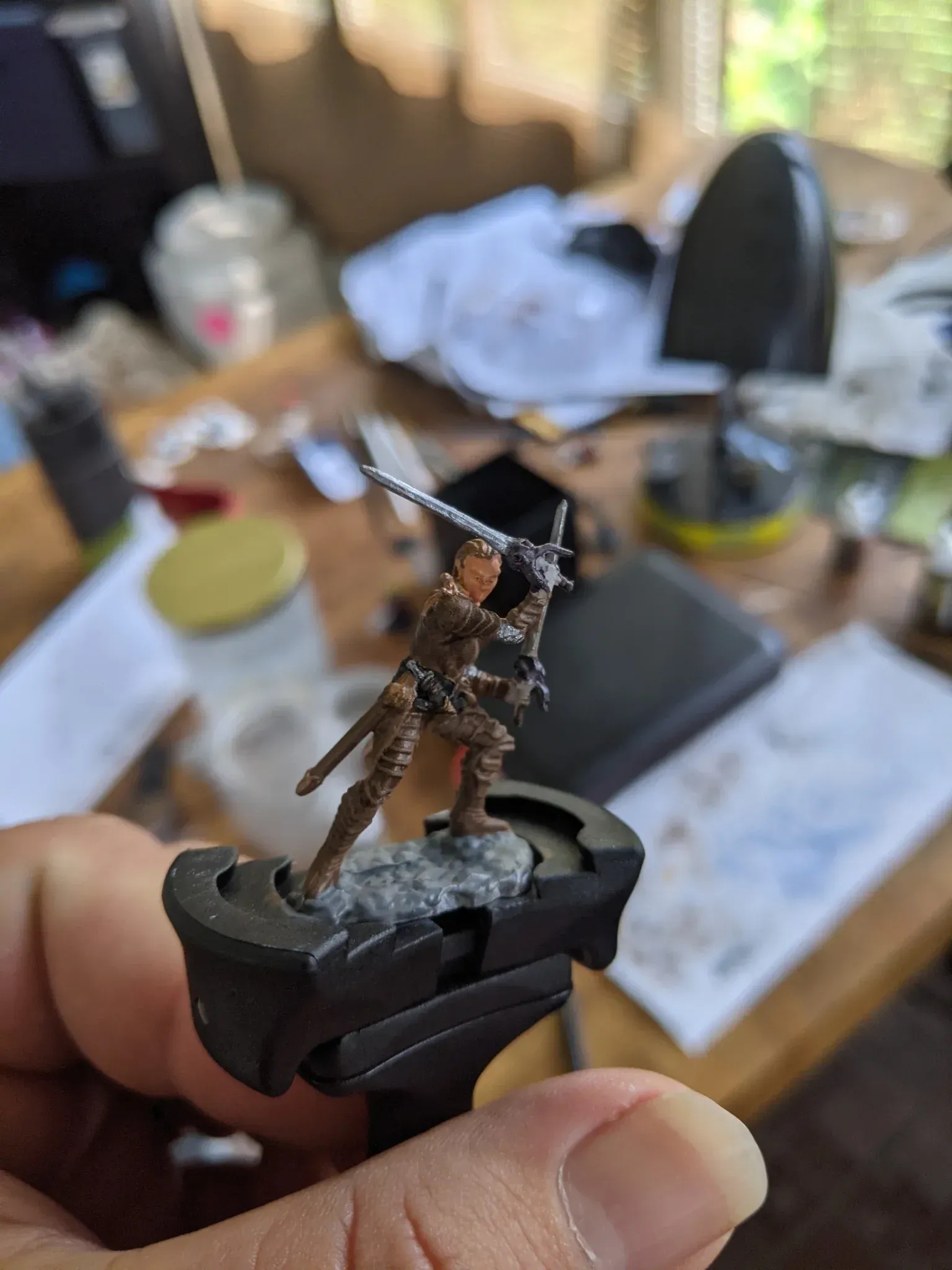 Miniature figure of a swordsman with brown armor, held in hand for painting, standing on a rocky base.