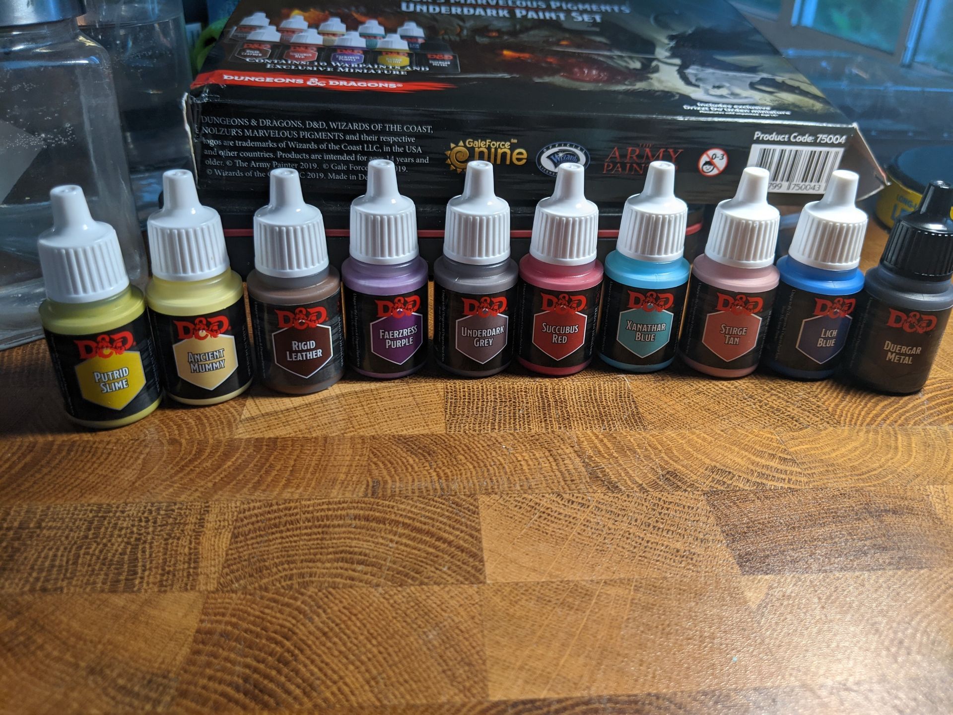 A row of D&D miniature paints: yellow, tan, brown, purple, maroon, red, teal, pink, blue, black, on a wooden surface.