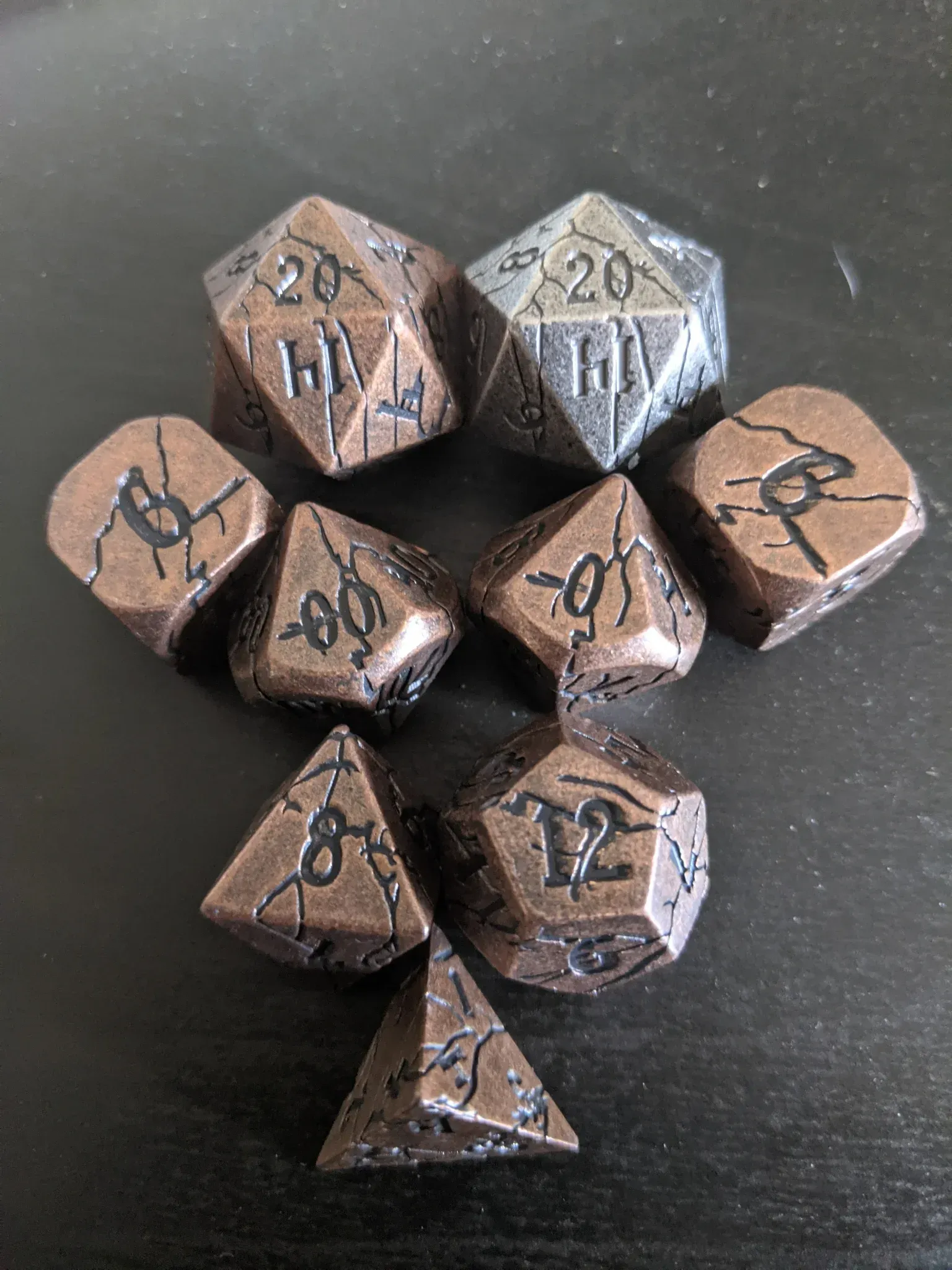 A set of metal dice in bronze and silver on a dark surface, used for tabletop games, including a d20.