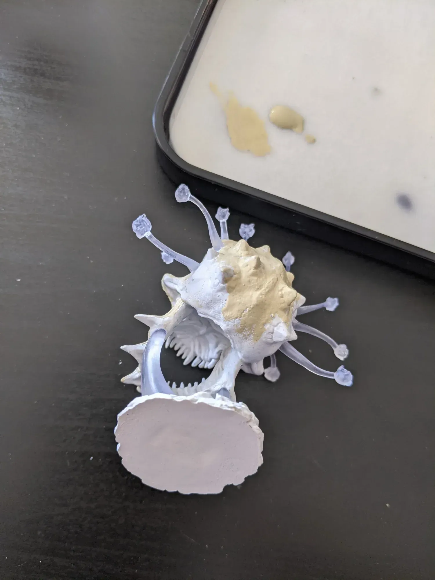 A partially painted miniature monster with spiky protrusions and teeth on a black surface, next to a paint palette.