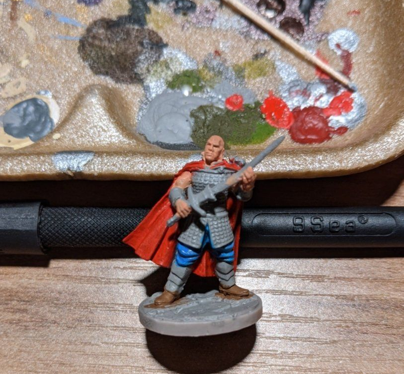 Miniature figure of a bald warrior with a red cape and sword, posed on a round base, next to a paint palette.