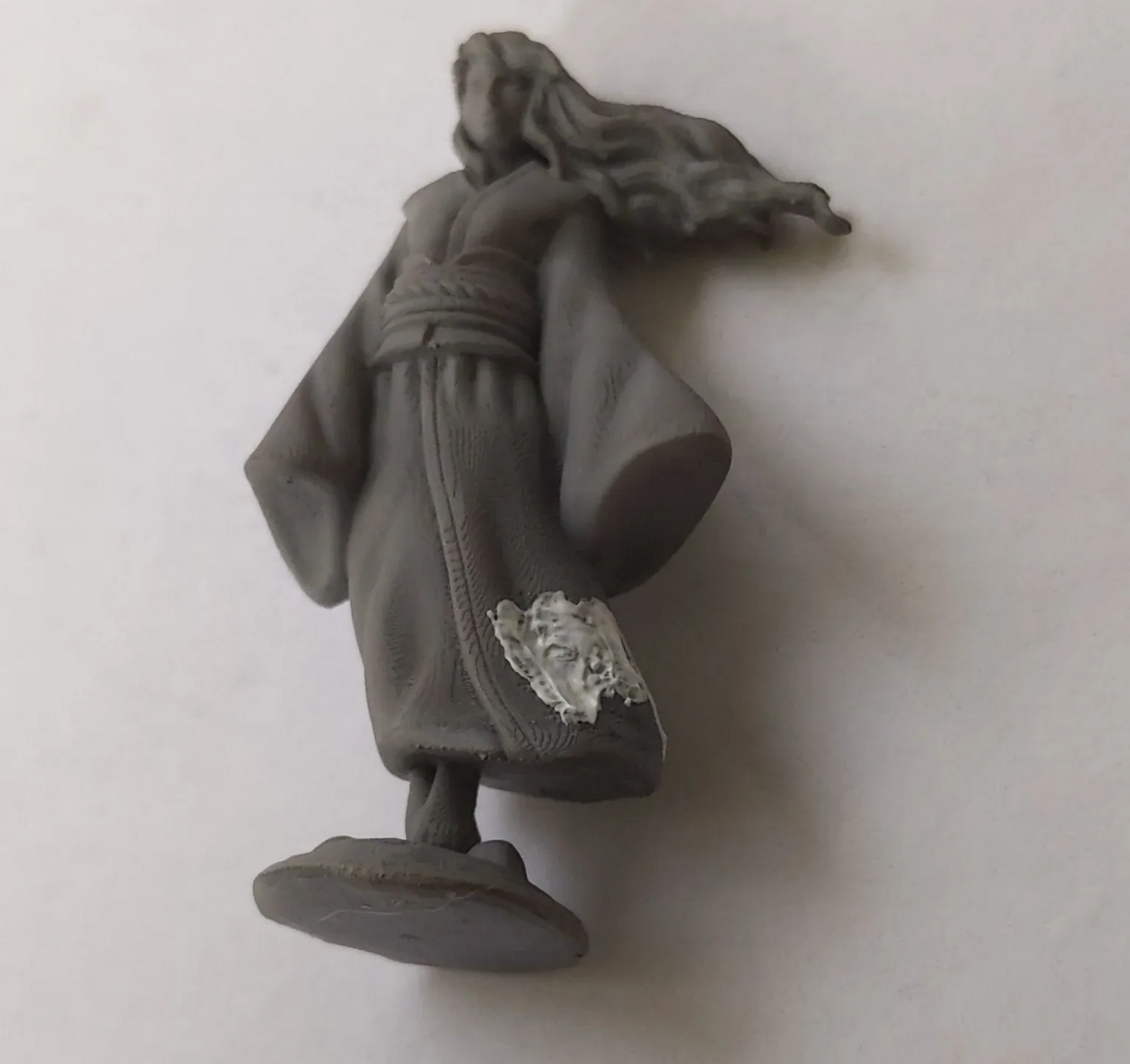 Gray miniature figure of a person in a robe, on a circular base, with long flowing hair.