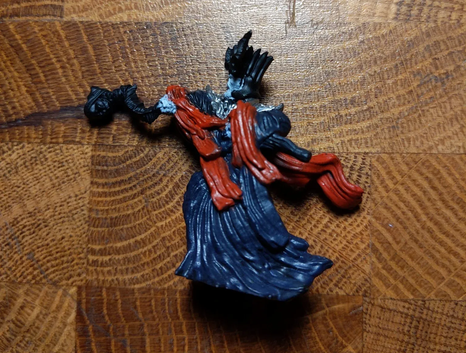 Miniature figure of a robed figure with red scarf, black arms, and dark crown, set on a wooden surface.