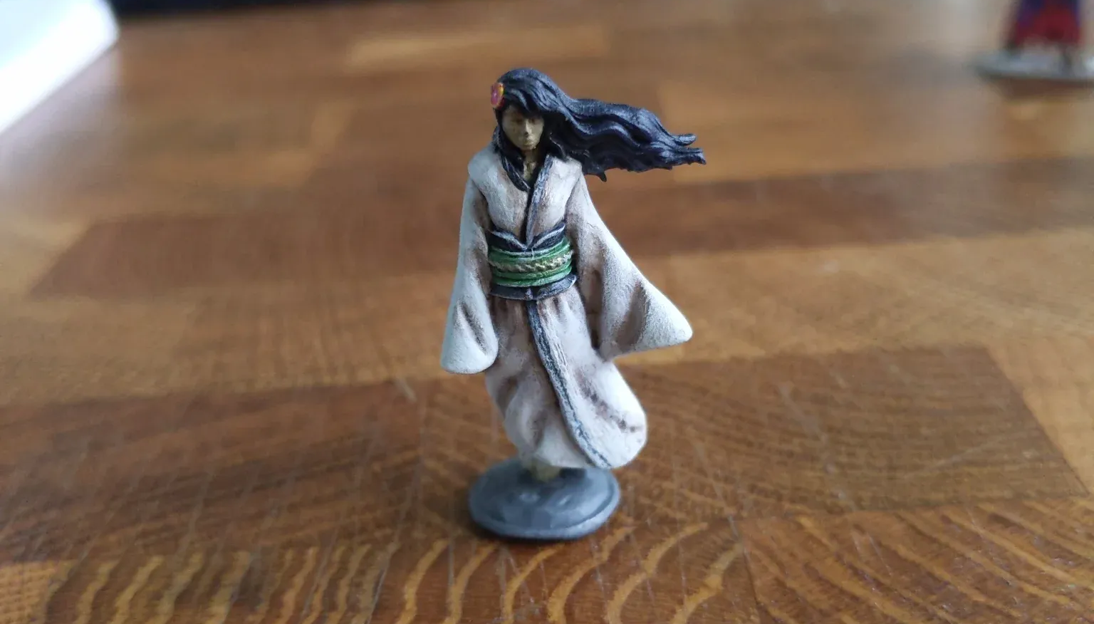 Miniature figure of a woman in a white robe, black hair, and a green sash, standing on a gray base.