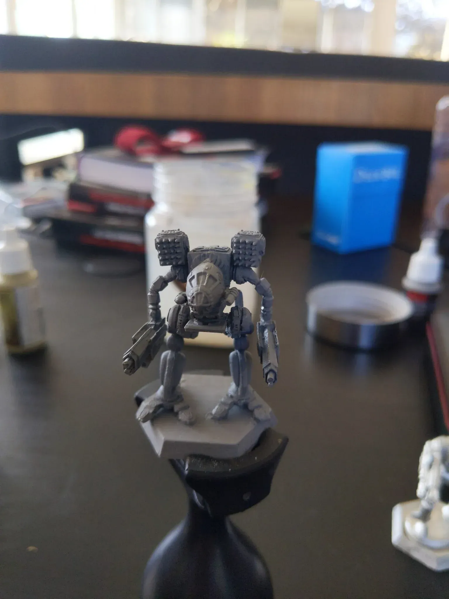 A miniature, gray BattleMech war machine model on a black stand, against a cluttered tabletop background.