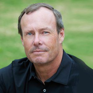 A man in a black shirt is sitting in the grass and looking at the camera.