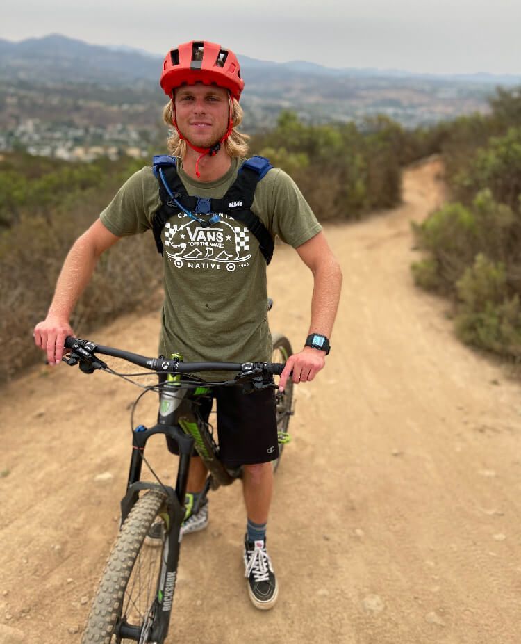 A man wearing a helmet is standing next to a bicycle on a dirt road.