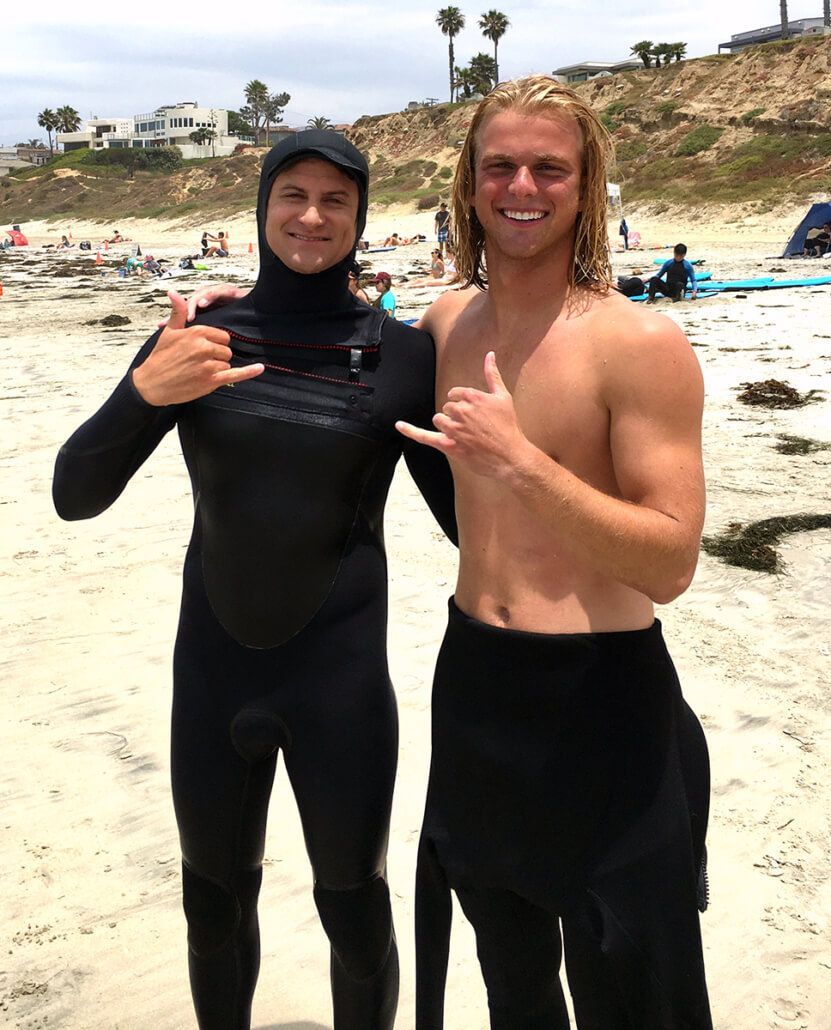 Two men in wet suits are posing for a picture on the beach.