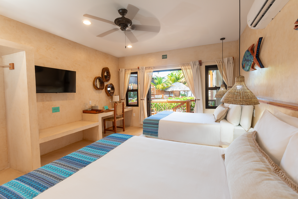 Bright hotel room with two beds, turquoise rug, wall-mounted TV, and balcony view through open doors