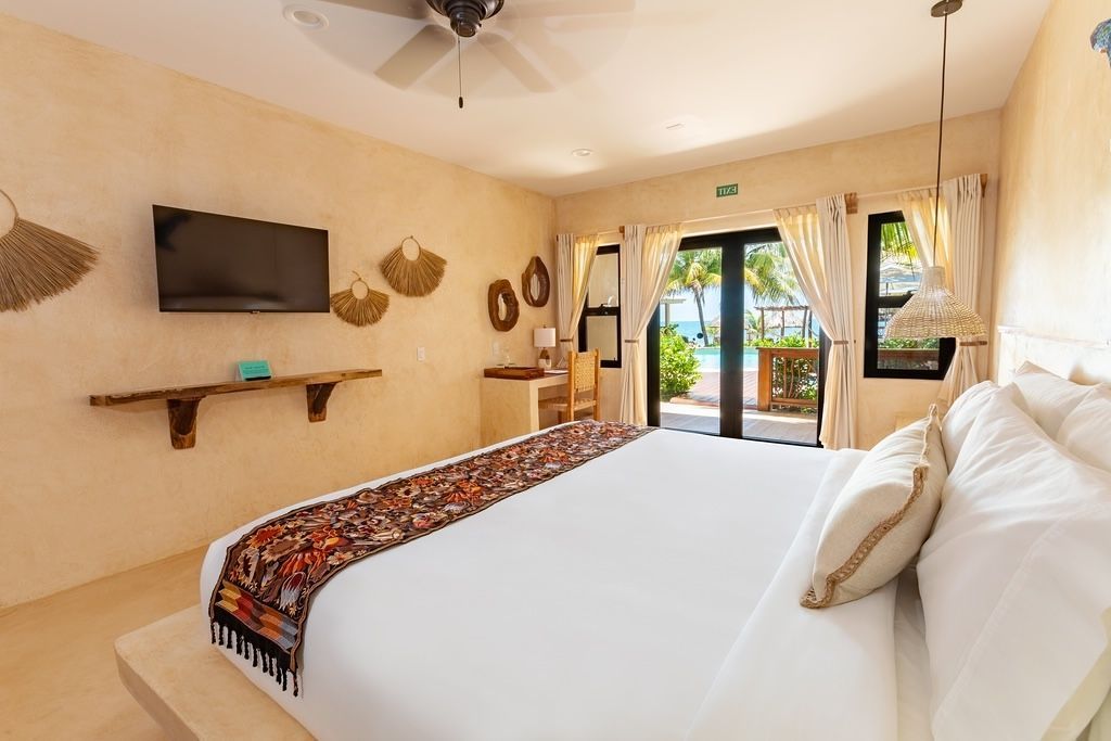 Bright hotel bedroom with king bed, wall-mounted TV, rustic decor, and balcony doors open to greenery
