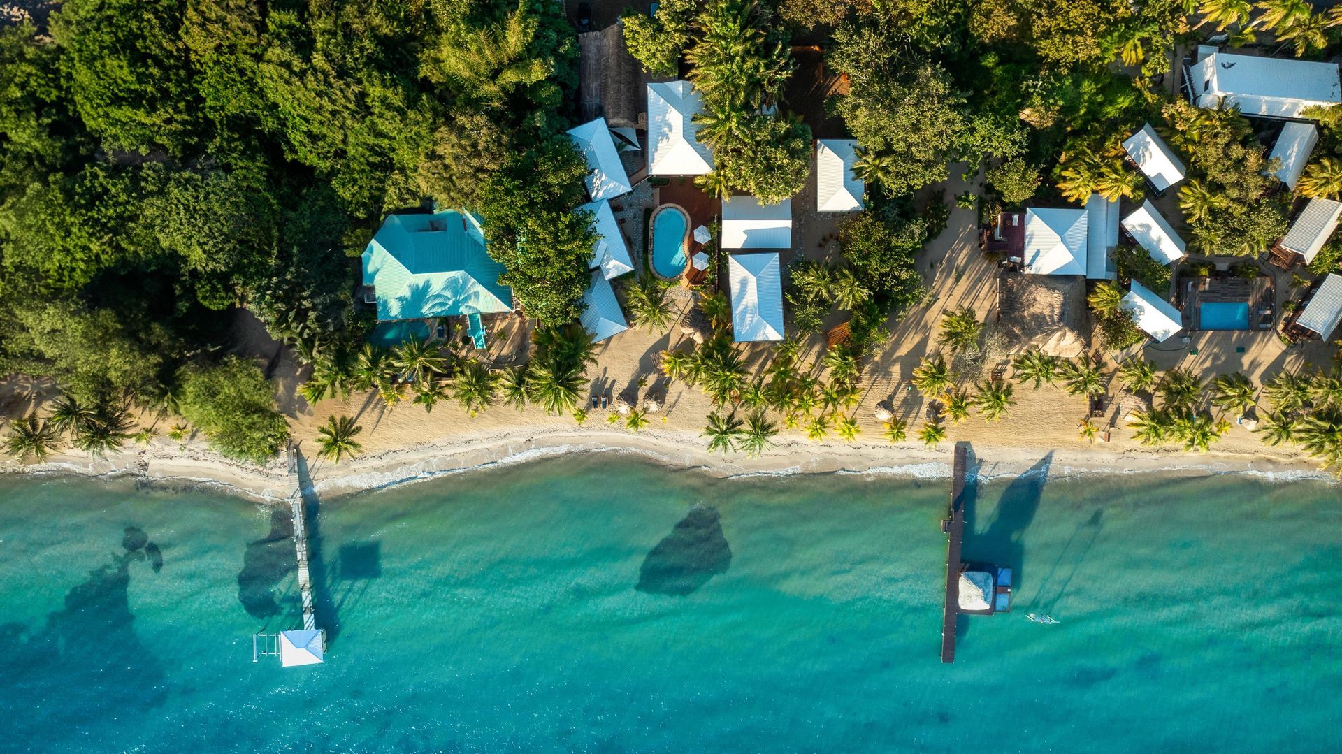 Aerial view of a tropical beach resort with white cabanas, blue water, sandy shore, and docked boats