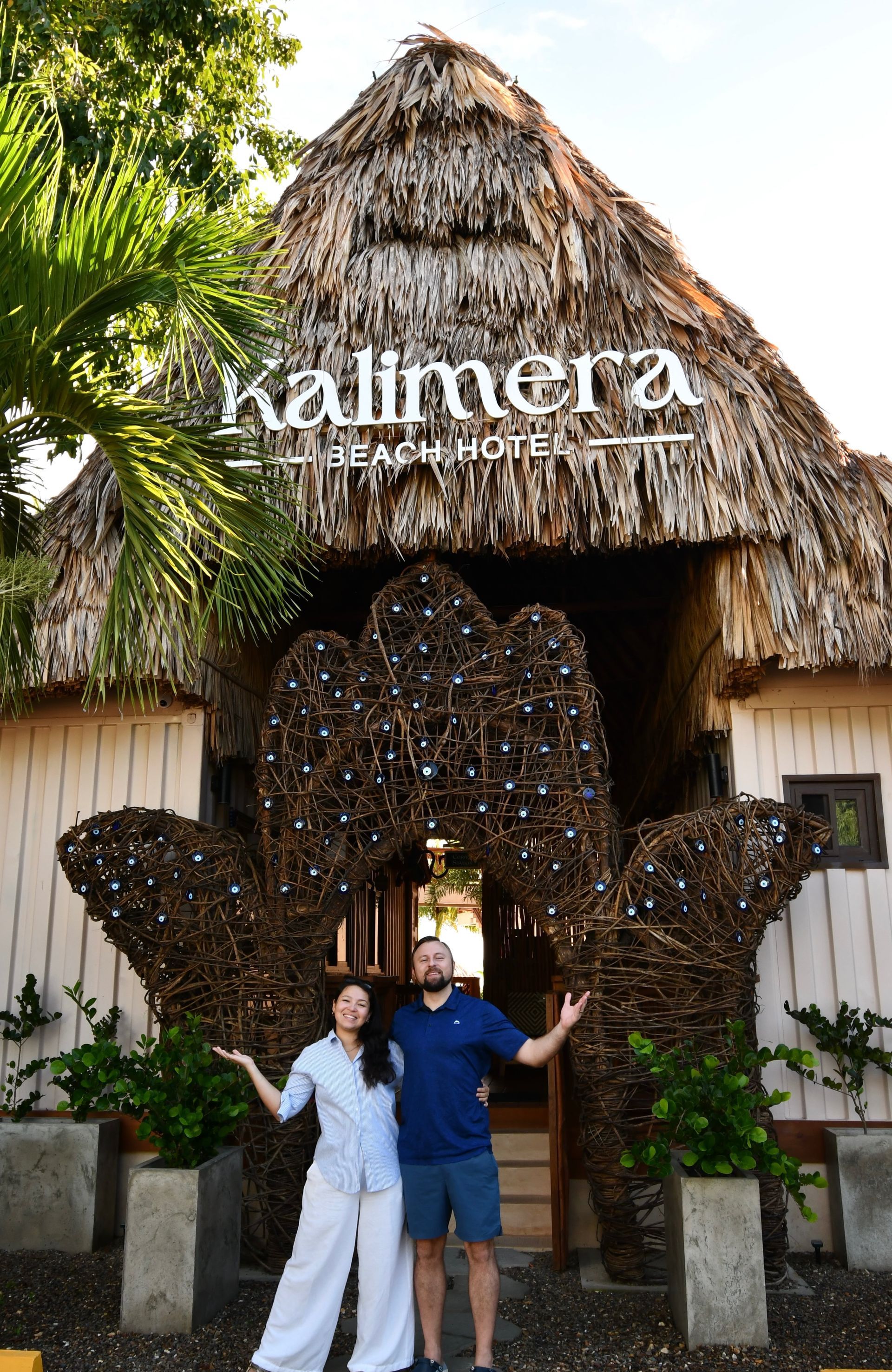 Two people posing under a thatched Kalimera sign at a tropical resort entrance