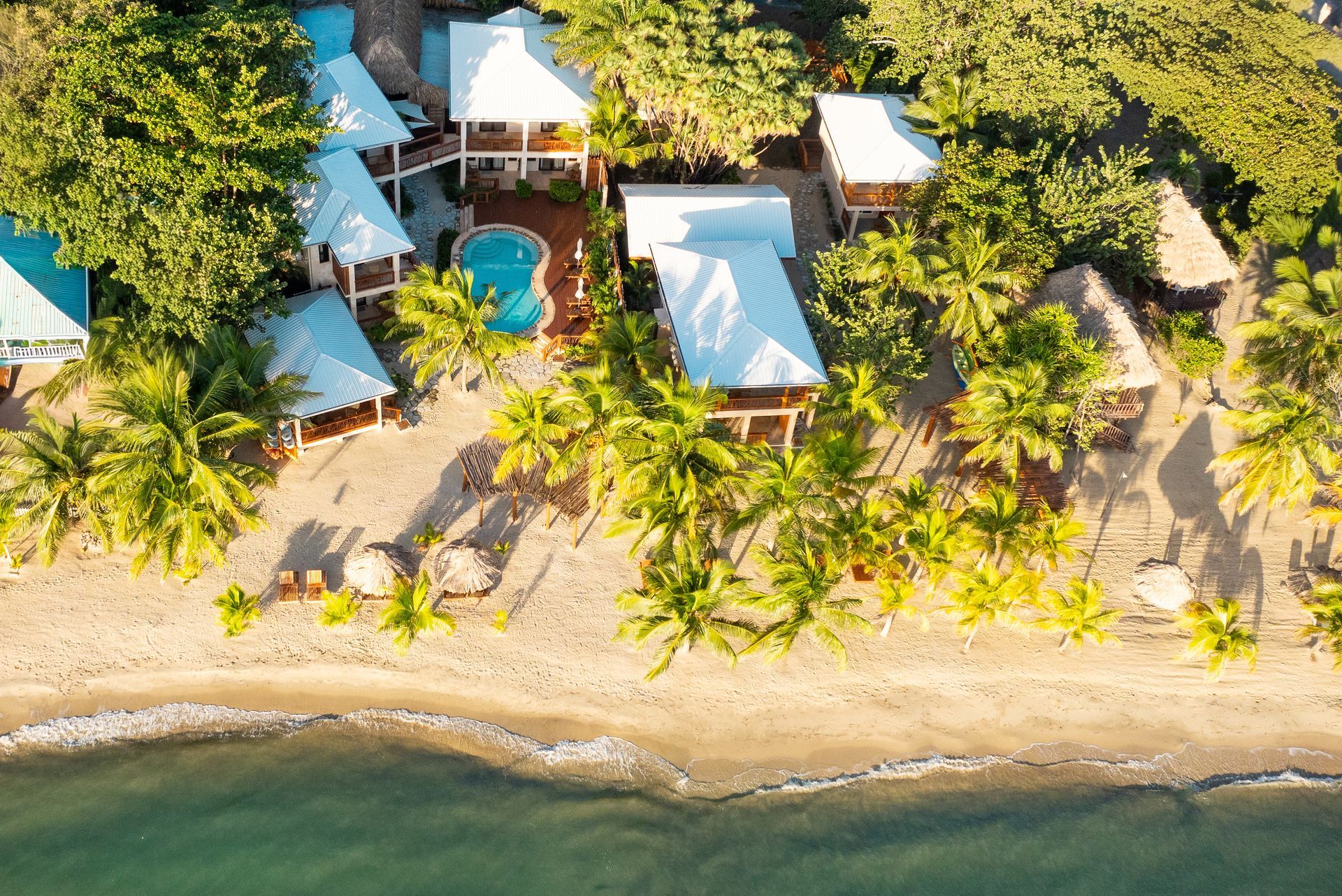 Aerial view of a tropical beachfront resort with blue-roofed bungalows, palm trees, and a sandy shore.